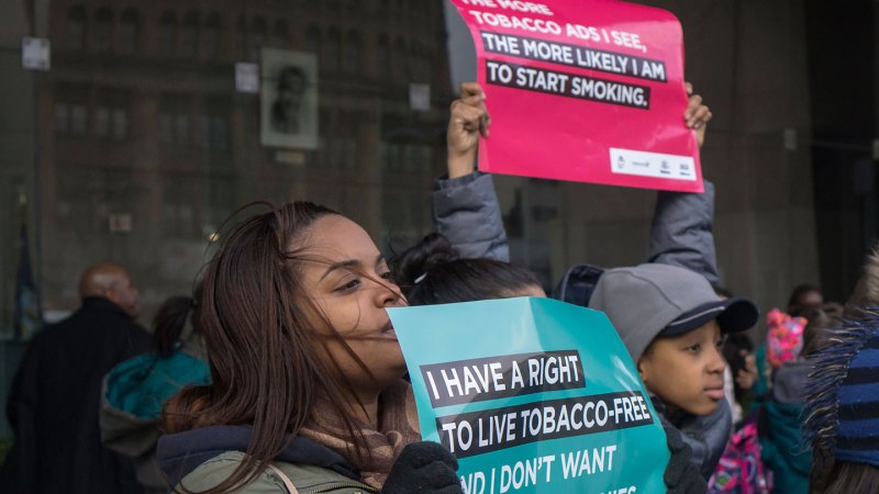 Two young people hold anti-tobacco signs at a a rally. The girl in front holds an teal-colored sign that reads, "I have a right to live tobacco-free." Another person holds a red sign that says, "The more tobacco ads I see, the more likely I am to start smoking."